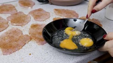 Dişi eller çiğ yumurtaları siyah bir kasede çatalla çırpar, yanında da baharatlı et pirzolası. Yakın çekim görüntüsü, ekmekli et yemeği hazırlamak için ev yapımı pişirme sürecini yakalar.
