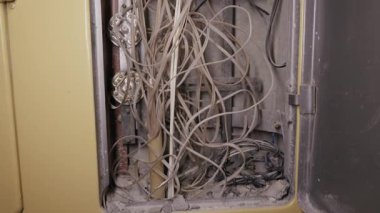 Eski elektrik ve telekomünikasyon kablolarından oluşan kaotik kütle, metal bir alet kutusunu dolduruyor. Kamera tozlu kabloları, paslı bağlantıları ve bakımsız altyapıyı gösteriyor..