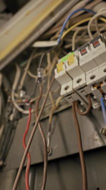 Eski devre kesiciler dağınık kablolarla çevrili tozlu metal bir panele monte edilir. Loş ışıklandırma kırmızı mavi ve kahverengi elektrik kablolarının kaotik düzenini vurgular.