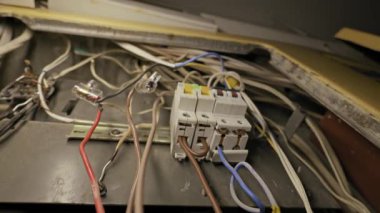 Metal bir bağlantı kutusundaki eski devre kesicilere bağlı dağınık elektrik kabloları. Kamera, loş ışıktaki kaotik kabloları ve tozlu bileşenleri ortaya çıkarmak için geri çekiliyor.