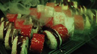Somon ve tobiko ile karışık maki suşi dürümleri karanlık bir tabakta dinleniyor. Yoğun kuru buz dumanı dramatik mavi ışık altında taze deniz ürünleri üzerinde dalgalanır..