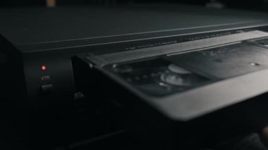 Erkek el, siyah VHS kaseti klasik VCR oynatıcı yuvasına yerleştirir. Yumuşak ışıklandırma, ahşap yüzeydeki eski elektronik ekipmanların dokusunu vurguluyor.