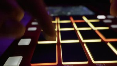 Bir müzisyenin elleri, kayıt sırasında bir davul makinesinin ışıklı pedlerine ritmik olarak dokunur. Müzik üretimi, gece hayatı yaratıcılığı ve modern ses teknolojisi kavramı.