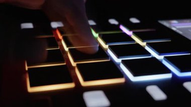 Bir müzisyen düşük ışıklı stüdyo ortamında bir MIDI kontrolörünün parlayan lastik pedlerine ritmik olarak dokunuyor. Elektronik müzik üretimi, dijital yaratıcılık ve modern ses teknolojisi kavramı.