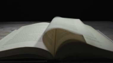 Açık bir kitap sayfasının, ahşap bir masanın üzerinde hızla dönen bir esintiye yakın çekimi. Öğrenme, hızlı okuma, hikaye anlatma, bilgi, tarih ve zamanın geçişi kavramı..