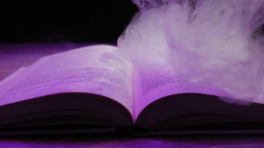 Açık bir ciltli kitap menekşe ışıklarının altında yatarken kalın duman sayfaların üzerinde kıvrılıyor. Büyü kavramı, fantezi hikayeleri, gizem ve cadılar bayramı atmosferi..