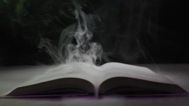 Açık bir ciltli kitap masanın üzerinde otururken renkli ışıkların altında sayfalarından yoğun duman yükseliyor. Sihir büyüleri, fantezi hikayeleri ve esrarengiz antik bilgi kavramı..
