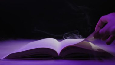 Kapkara dumanla çevrili açık bir kitabın sayfasını çeviriyor. Sihir büyüleri, fantezi hikayeleri, gizem ve yasak bilgi kavramı..