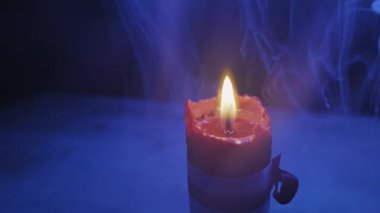 Karanlıkta alevin etrafında dönen kalın mavi dumanlı, yanan kırmızı bir mumun yakın çekimi. Gizem kavramı, ruhanilik, sihir ritüelleri ve ürkütücü Cadılar Bayramı atmosferi..