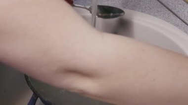 Kırmızı ojeli bir kadın kaşık ve metal bir kâse ile mutfak lavabosunda akan suyun altında sünger yıkıyor. Ev işleri, ev hayatı, günlük rutin ve kişisel sorumluluk kavramı.