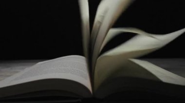 Açık kitap sayfalarının yakın plan çekimleri siyah arka plana karşı rüzgarda savruluyor. Bilgi, eğitim, hikaye anlatma, tarih ve zaman geçişi kavramı.