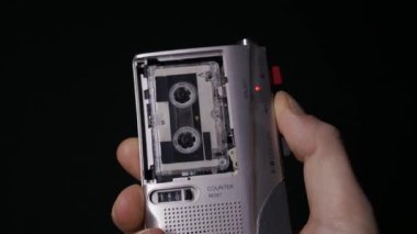 Bir el siyah arkaplana karşı gümüş bir mikrokaset kaydedici tutuyor ve kaseti başlatmak için oynatmaya basıyor. Retro teknoloji kavramı, araştırmacı gazetecilik ve analog ses kanıtı.