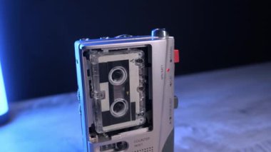 Gümüş renkli mikro kasetçalar, ahşap bir masanın neon ışıklarının değişmesine karşı dönen makaralı sesleri kaydeder. Casusluk anlayışı, retro teknoloji, 80 'lerin nostaljisi ve lofi ses kaydı..