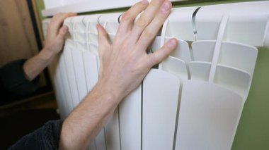 Bir adamın eli, ısıyı kontrol etmek için yeşil bir duvara monte edilmiş beyaz alüminyum radyatöre dokunuyor. Kışın ısıtma mevsimi kavramı, pahalı enerji faturaları ve ev sıcaklığının korunması..