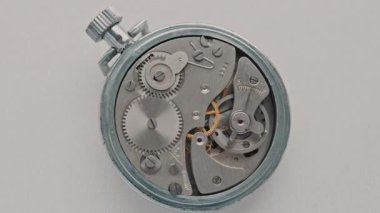 Yukarıdan aşağıya bakan bir makro görüntü beyaz üzerinde dönen gümüş bir kronometrenin karmaşık iç dişlilerini gösterir. Hassas mühendislik, zaman yönetimi ve karmaşık mekanik sistemler kavramı.