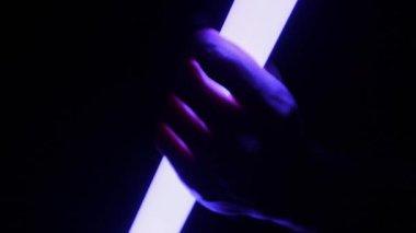 Bir el, parlak bir LED tüp ışığını siyah bir arkaplanla kavrar, maviden mora ve turuncuya renk değiştirir. Bu kavram yaratıcı aydınlatma ve gelecekteki estetiği gösteriyor..