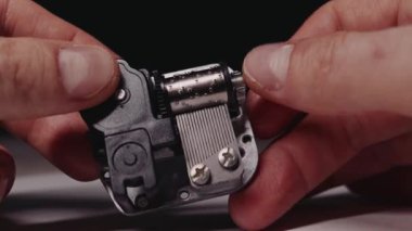 Hands hold a small metal music box mechanism and turn the crank to play a melody. The rotating cylinder plucks the comb. Concept highlights nostalgia and simple mechanical engineering.