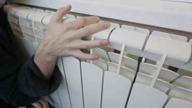 Bir adamın eli odadaki sıcaklığı kontrol etmek için merkezi ısıtma radyatörünün beyaz kısımlarına dokunuyor. Ev konforu, artan enerji maliyetleri ve kış ısıtma mevsimi kavramı.