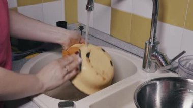 Kırmızı ojeli bir kadın, akan suyun altında kirli sarı bir kâseyi süngerle yıkıyor. Ev işleri, ev işleri, temizlik ve günlük rutin işler kavramı.
