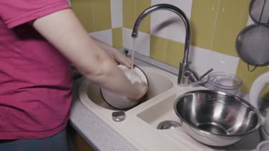 Tanınmayan bir kadın mutfak lavabosunda duruyor ve krom musluktan akan suyun altında süngerle dikkatlice bir kase yıkıyor. Günlük ev işleri, ev işleri ve hijyen kavramı.