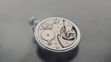 Bir makro çekim, karanlık yansıtıcı bir masada, mekanik bir cep saatinin açıkta kalan dişlilerinin yörüngesinde döner. Karmaşık mühendislik kavramı, zaman yönetimi ve antika burçların hassasiyeti..