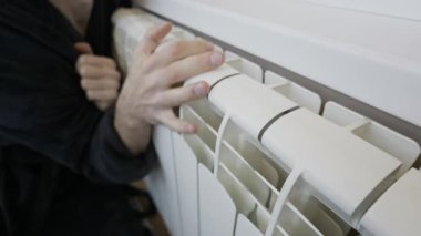 Odanın içindeki sıcaklığı hissetmek için bir adamın beyaz metal bir radyatöre dokunuşunun yakın plan görüntüsü. Isınma mevsimi kavramı, artan enerji maliyetleri ve soğuk kış günlerinde ısınma kavramı.