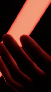Bir el, parıldayan kırmızı bir LED ışık çubuğunu nazikçe kavrar ve zifiri karanlığa karşı derin gölgeler yaratır. Fütürist teknoloji, gizem ve soyut aydınlatma efektleri kavramı.
