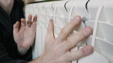 Male hands touch a white central heating radiator to check the temperature and warm up frozen palms inside a cold apartment. This concept highlights home energy efficiency and heating costs.