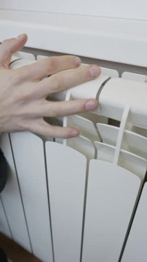 Bir adamın eli soğuk, beyaz bir radyatöre dokunur ve dikey çekimde sıcaklığı kontrol eder. Isınma mevsimi kavramı, artan enerji maliyetleri ve ev faturaları.