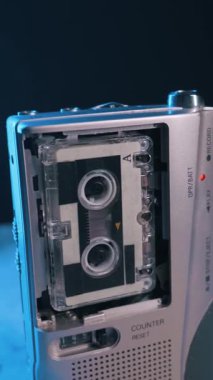 Gümüş renkli taşınabilir bir mikrokaset kaydedici, renk değiştiren ışıkların arka planına karşı dönen makaralarla çalışır. Retro teknoloji kavramı, lofi estetik ve nostaljik ses izleme..