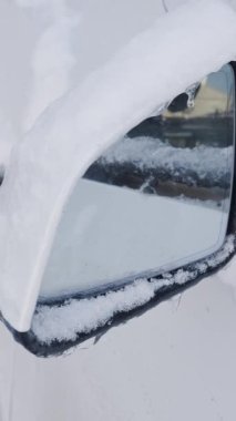 Yakın plan dikey çekimde yoğun kar ve buz saçakları ile kaplı beyaz bir araba yan aynası görülüyor. Dondurucu hava koşulları, kış sürüş güvenliği ve soğuk mevsimsel iklim kavramı.