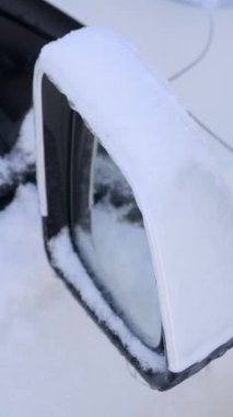 Yakın plan dikey bir karla kaplı beyaz bir araç yan aynası görüntülenir. Kış sürüş zorlukları, soğuk hava bakımı ve mevsimlik araba bakımı kavramı.