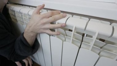 Bir adam ısıyı kontrol etmek için beyaz merkezi ısıtma kaloriferine uzanır ya da donan ellerini ısıtır. Enerji krizi kavramı, artan ısıtma maliyetleri ve kış boyunca ev konforu