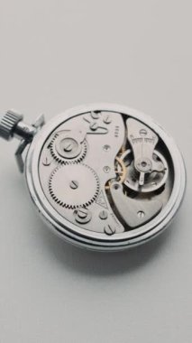 Bir makro görüş beyaz bir yüzey üzerinde dönen klasik gümüş kronometre mekanizmasının karmaşık dişlilerini gösterir. Hassas mühendislik, zaman geçirme ve mekanik sanat kavramı.