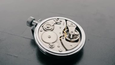 Eski bir mekanik kronometrenin gümüş iç dişlileri ve yayları karanlık bir masanın üzerinde duruyor. Hassas mühendislik kavramı, zamansız işçilik ve karmaşık mekanizma..