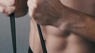 Gövde ve karın kaslarını tanımlayan tişörtsüz bir adamın önden yakın çekimi. Siyahi direniş bandını germek için iki elini de kullanıyor. Güç kazanmak için dinamik göğüs egzersizi yapıyor..
