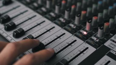 Açılı yakın çekim prodüktörünün eli ses panosundaki iki fadarı aynı anda ayarlıyor. Çekimde serin ışıklandırma, sığ alan derinliği ve stüdyoda birden fazla aydınlatma düğmesi yer alıyor..