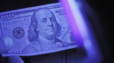 Ultraviyole ışık taraması yaparken yüz dolarlık banknotu tutan bir elin makro görüntüsü. Işık gizli güvenlik ipliğini ve florasan özellikleri ortaya çıkarıyor..