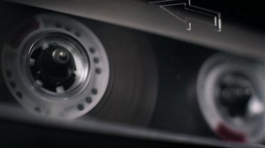 Dönen beyaz bir makarayla çalan şeffaf bir ses kasetinin açılı makro görüntüsü. Kabartmalı ok işareti koyu bant destesinin içindeki kabukta görünür..