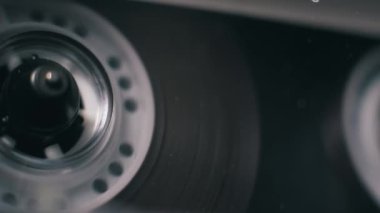 Kırmızı işaretlerle oynayan şeffaf bir ses kasetinin açılı yakın çekim görüntüsü. Sol makara ve manyetik film karanlık ve kasvetli bir ortamda döner..