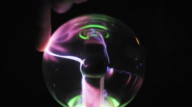 Bir parmağın yakın çekimi cam plazma küreye dokunuyor ve tek bir mor elektrik ışını parmak izini takip ediyor. Koyu arkaplan, bir bilim ortamında neon yeşil yansımalar içerir.