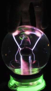 Bir insan elinin dikey görüntüsü plazma küre cam kubbe üzerinde hareket ediyor. Yeşil ışık küreyi aydınlatırken, çoklu beyaz ve mor elektrik akımları parmak uçlarını takip eder..