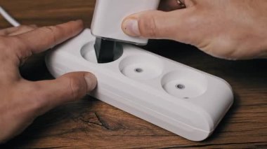 Eller, beyaz şarj aleti ve siyah topraklama kablosu da dahil olmak üzere birden fazla aleti beyaz bir soket güç çubuğuna takar. Tahta masadaki düzenek güç tüketimini ve kullanımını gösteriyor.