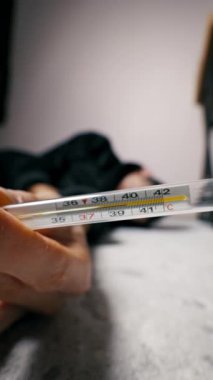 Ellerinde cıva termometresi var. Ateşi 40 santigrat derece. Hasta biri arka planda bulanık yatıyor. Yakın çekim, hastalığın ciddiyetini vurgular..