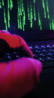 Eldivenli eller hızlı bir şekilde arka planda dizüstü bilgisayarda klavye kullanıyor. Kırmızı ve mavi neon ışıklı karanlık bir odada ekranda yeşil matris tarzı parşömenler.