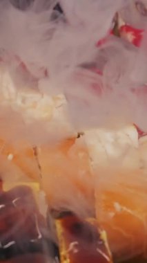 Somon, yılan balığı ve kırmızı yumurta da dahil olmak üzere çeşitli suşi ruloları karanlık bir konteynırda otururlar. Kalın beyaz duman taze Japon deniz ürünlerinin üzerinde kıvrılarak dramatik bir sunum oluşturur..