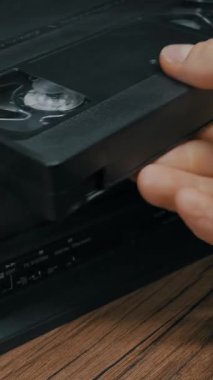 Yakın plan dikey çekimde, bir elin tahta bir masanın üzerindeki bir video oynatıcıya VHS bandı soktuğunu görüyoruz. 90 'ların nostaljisi, analog teknolojisi ve retro ev eğlencesi kavramı.