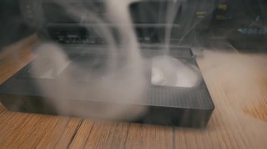 Siyah plastik VHS kaset kaseti VCR oynatıcının önündeki ahşap masada duruyor. Yoğun beyaz duman, yavaş yavaş havaya karışmadan önce retro ortam nesnesinin etrafında dönüyor.