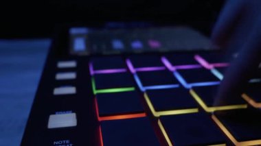 Bir müzisyen, bir midi kontrolörünün renkli LED pedlerine vurarak ritim yaratır. Elektronik müzik üretimi, yaratıcı stüdyo iş akışı ve modern ses teknolojisi kavramı.