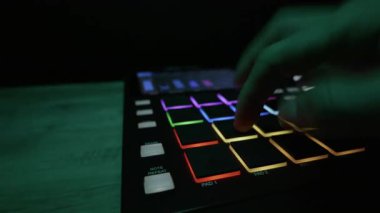 Bir müzisyenin eli, karanlık bir odada renkli bir davul makinesinin LED pedlerine ritmik vuruşlar yapar. Elektronik müzik üretimi, yaratıcı DJ yaşam tarzı ve stüdyo teknolojisi kavramı.